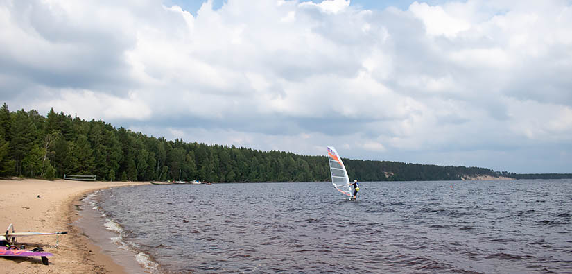 Leijasurfaaja järvellä lähellä hiekkarantaa. Rannalla on toinen surffilauta ja kauempana veneitä laiturissa.