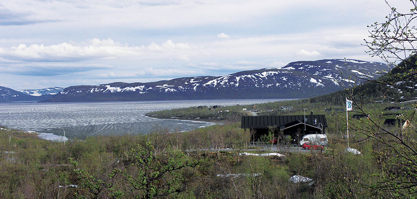 Järven rannalla on rakennuksia ja lipputanko. Kauempana on tuntureita, joiden rinteillä on laikuttain lunta.