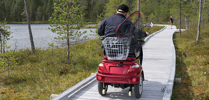 Mies ajaa sähköisellä pyörätuolilla pitkospuita pitkin. Mopon kyydissä on kalahaavi.