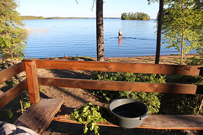 Nainen on menossa uimaan. Etualalla ovat saunan kaiteet, pesuvati ja vasta. Järvellä näkyy pieni saari.