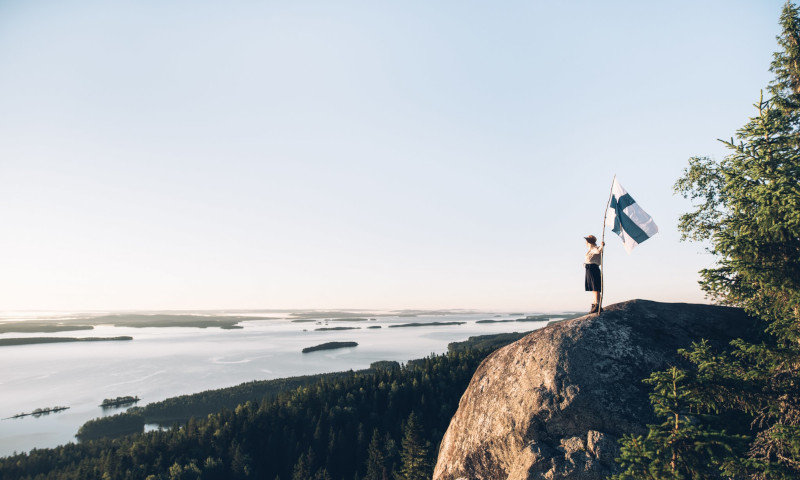 Nainen seisoo Suomen lipun kanssa Kolin maisemassa.