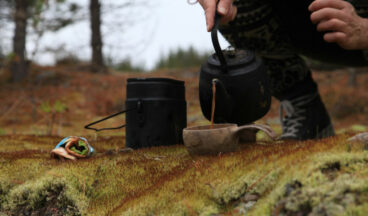 A hiker is pouring coffee brewed over a campfire into a wooden cup.