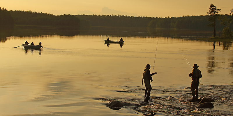 Kalastajia järven rannalla illalla ja soutajia soutuveneissä taustalla.