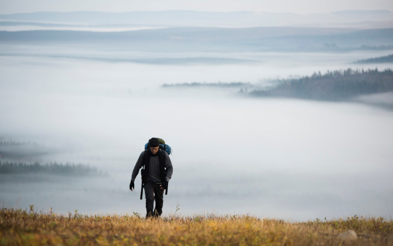 En man klättrar uppför en skogklädd sluttning med ett dimmigt skogslandskap i bakgrunden.