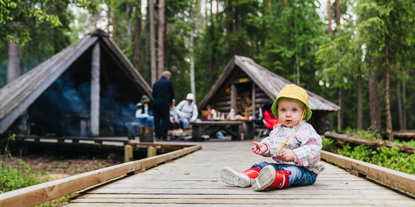 Lapsi istuu lankuista tehdyllä kulkutiellä, taustalla kaksi hirsilaavua ja eväiden syöjiä metsässä Napapiirin retkilyalueella Rovaniemellä.. Kuva Jaakko Posti.