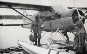 En man utrustad för vandring hoppar i land från ett pontonflygplan, svartvit bild.