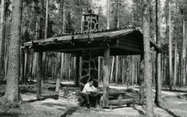 En man sitter vid ett bord vid en takförsedd eldplats i Petkeljärvi nationalpark, svartvit bild.