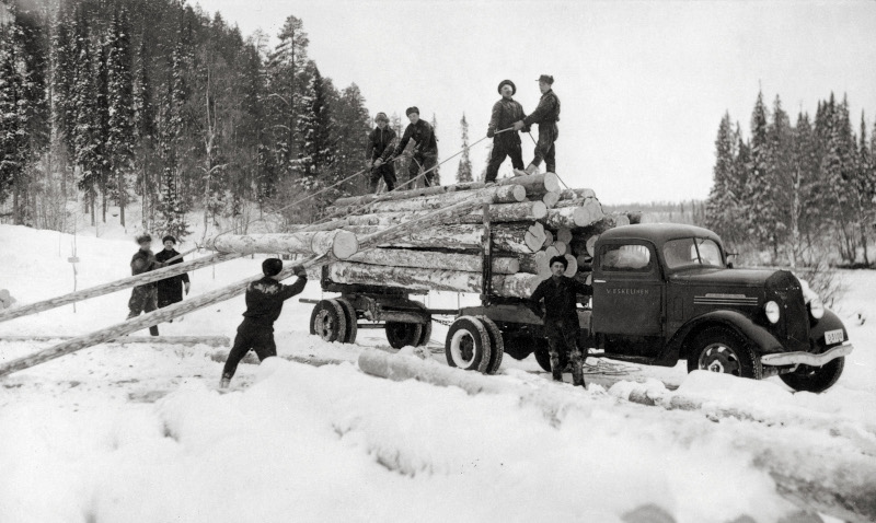 Män lastar timmerstockar på en bil vid en snöig skogsväg, svartvit bild.