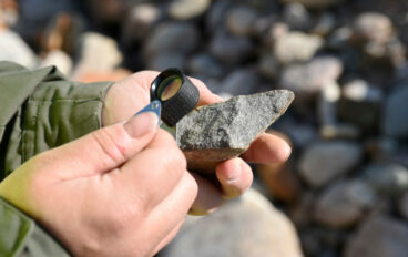 Henkilö tutkii kivenpalaa suurennuslasilla, lähikuva käsistä.