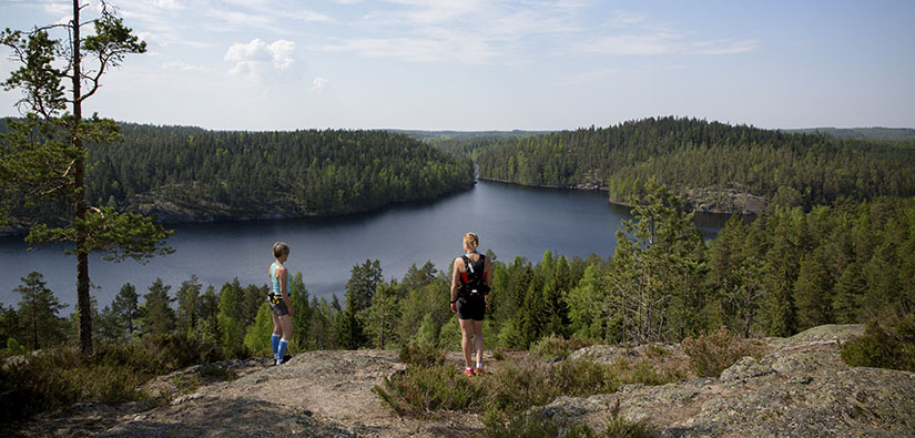 Kaksi juoksijaa katselee maisemia korkealla kalliolla. Alhaalla näkyy järvi ja metsää.