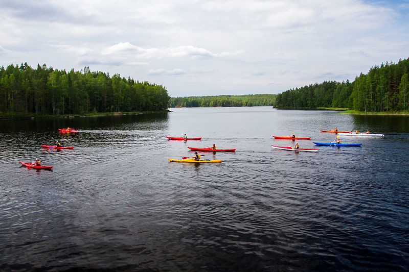 A dozen canoes are travelling along a calm lake. There are islands in the background. 