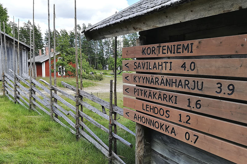 Perinteinen pihapiiri, jossa punamullalla maalattu asuinrakennus, riukuaita ja kaksi harmaata talousrakennusta, joista toisen seinässä on reittiopasteita.