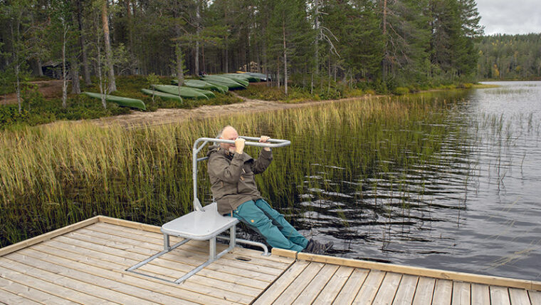 Ihminen istuu telineessä, johon voi laiturilla siirtyä pyörätuolista ja laskeutua laiturin vieressä olevaan kanoottiin.