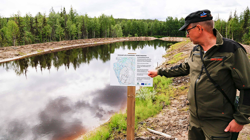 Mies osoittaa infotaulua vesialtaan rannalla, ympärillä metsää.