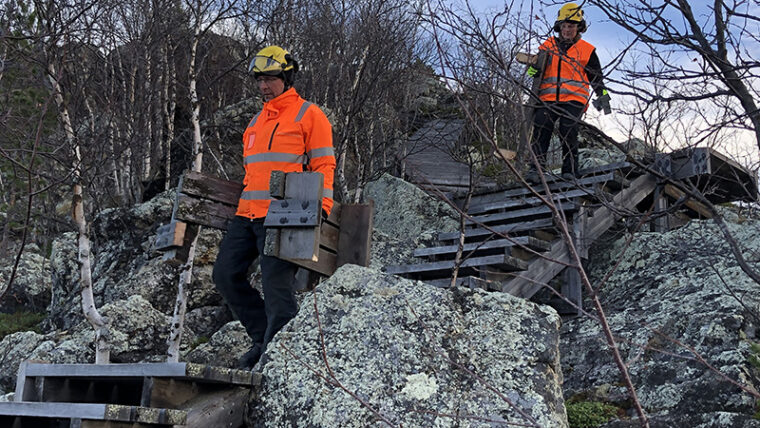 Kaksi ihmistä turvakypärä päässä laskeutuu alas puisia portaita kivikkoisella mäellä ja kantaa irrotettuja porraslautoja mukanaan.