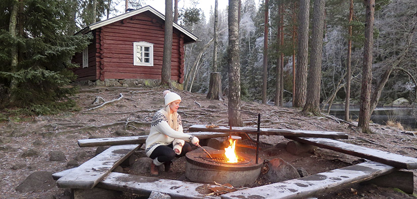 Retkeilijä istuu taukopaikan nuotiolla. Taustalla on punamultainen rakennus.