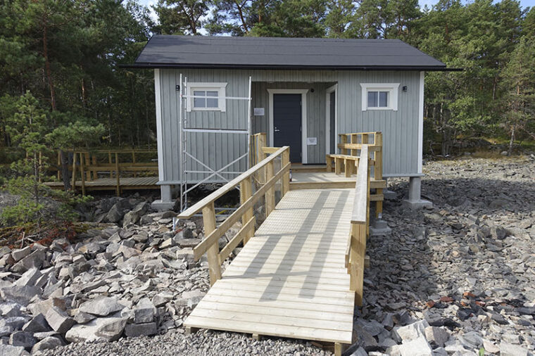 Small grey building with wooden ramp and handrails leading to it.