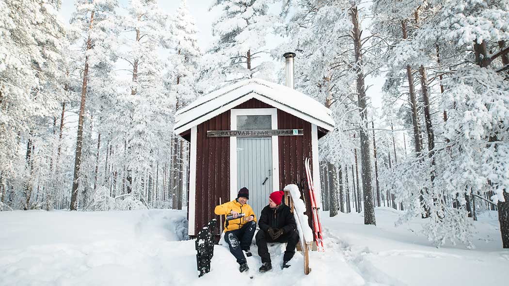 Kaksi retkeilijää istuu kahvitauolla pienen rakennuksen lumisilla portailla. Taustalla on luminen metsä.