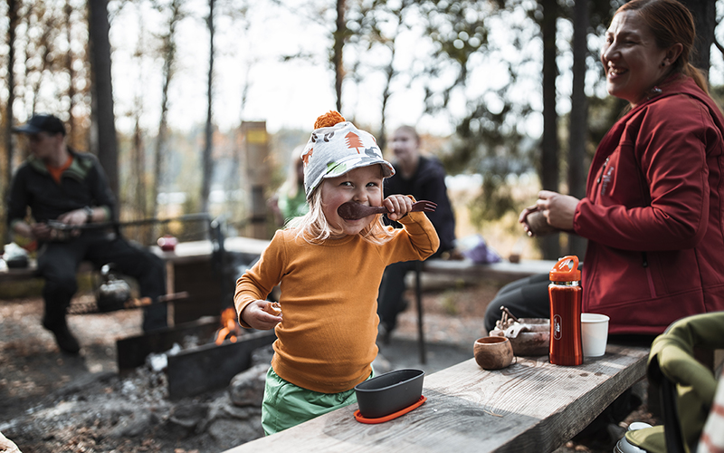 A smiling child eating with a wooden spoon outdoors. Other people and a fireplace in the background.