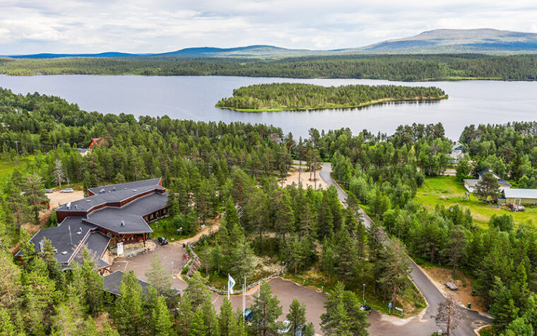 Etualalla puiden keskellä rakennus ja teitä ilmasta kuvattuna, taustalla järvi ja tuntureita.