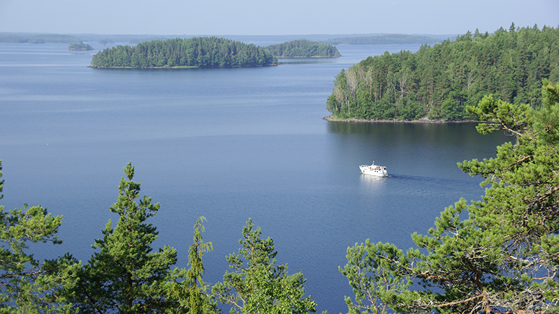 Puunlatvojen tasalta avautuva kesäinen järvimaisema, missä saaria ja laiva.