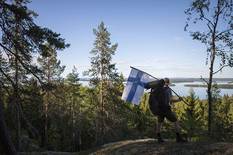 Retkeilijä seisoo vaaran laella katsomassa järvimaisemaa. Retkeilijällä on kädessään kookas Suomen lippu ja selässään rinkka.