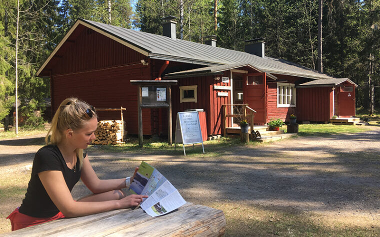 Ihminen istuu kseäpäivänä ulkona pöydän ääressä tutkimassa esitettä, takana hirsirakennus, jonka ovi auki ja vieressä kahvilan kyltti.