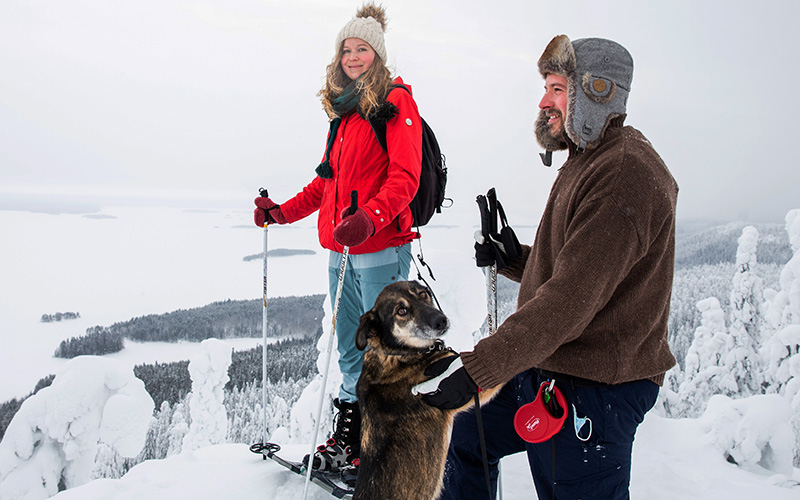 Kaksi ihmistä ja koira korkean vaaran huipulla talvella, maasto paksun lumen peitossa.