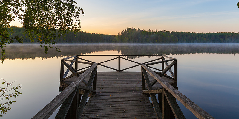 Kaiteilla varustettu puulaituri tyynen järven rannalla kesäauringon laskiessa.