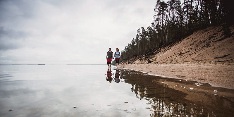 En man och en kvinna går i strandvattnet. En tallskog växer på sjön.