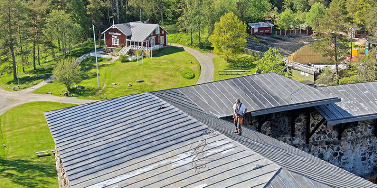 En människa står på stenslottets plåttak. Bakom slottet träbyggnader och gräsfält.