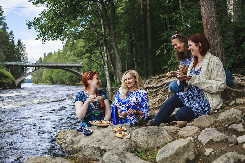 Neljä naista istuu virtaavan joen rantakivillä juomassa kahvia ja syömässä mustikkapiirakkaa. Kaikilla naisilla on sinivalkoiset vaatteet ja he nauravat.