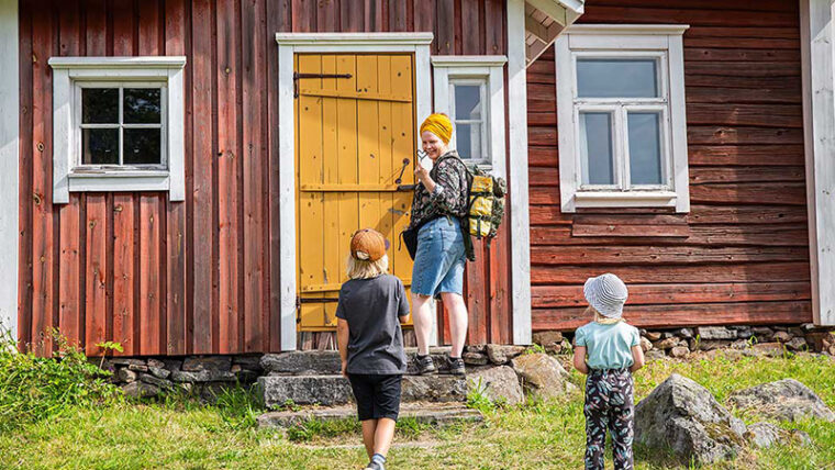 Nainen ja kaksi lasta vanhan puurakennuksen portailla, naisella avain oveen.