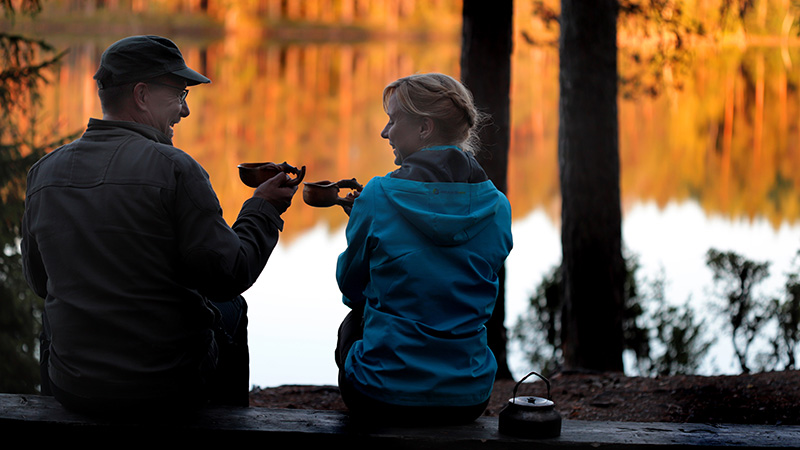 A man and a woman are sitting on a bench and drinking coffee by the lake.