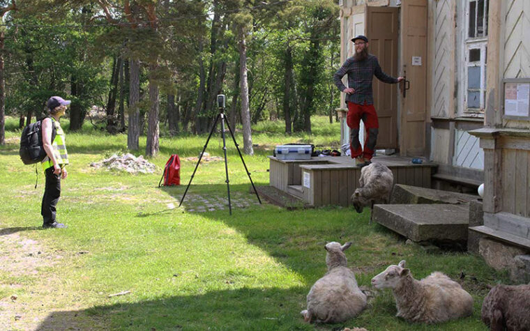 Ihmisiä ja lampaita vanhan puurakennuksen edustalla, taustalla mäntymetsää.