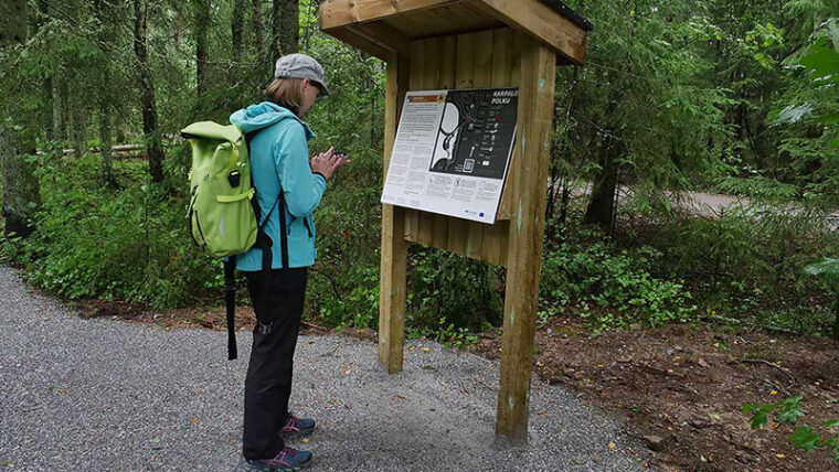 Ihminen reppu selässä sorapolulla metsässä infotaulun edessä.