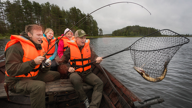 Young men and women sitting in a boat fishing. The men have lifted the fish into the net.