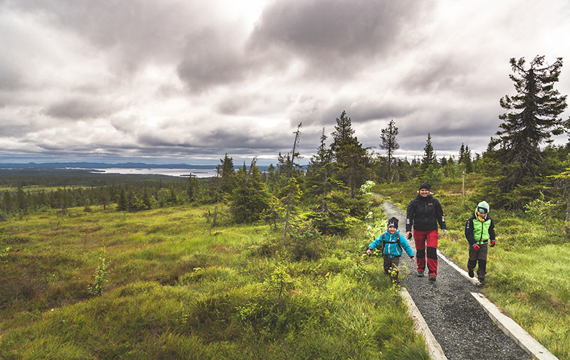 En kvinna går med två barn på en grusväg i ett öppet bergslandskap. Det finns plankor vid banans kanter. Bakom det finns en sjö och en granskog.