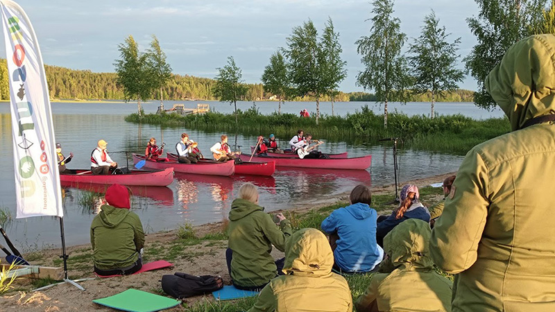 Hiekkarannalla istuu ja seisoo ihmisiä katsomassa vesirajaan, missä vierekkäin kanootteja ja niissä kitaran ja puhaltimen soittajia soittamassa.