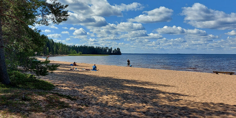 Suuren järven hiekkarannalla muutamia ihmisiä kesäpäivänä, rannoilla havumetsää.