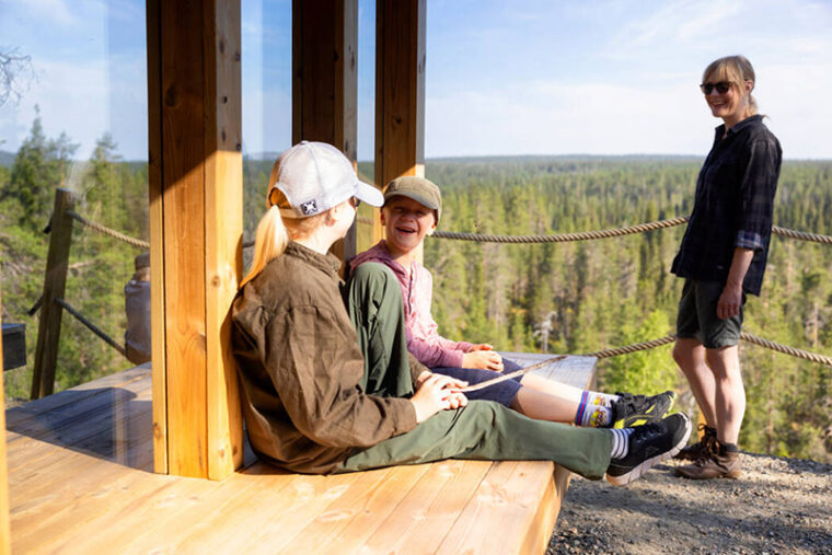 Kaksi hymyilevää lasta istuu rakennuksen penkillä. Taustalla seisoo hymyilevä nainen. Kuva on otettu kesällä.