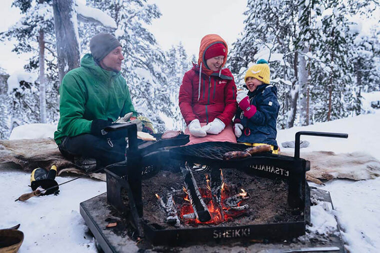 Kaksi aikuista ja lapsi istuvat porontaljojen päällä nuotiopaikalla talvella.