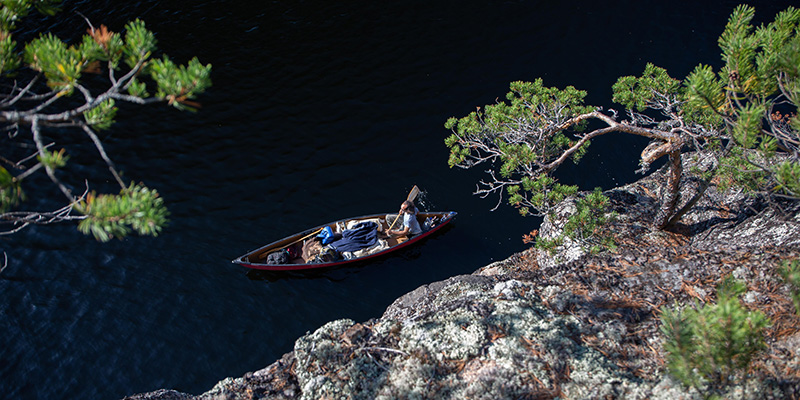 Ylhäältä kallion reunalta kurkistus alas järvelle, missä meloja avokanootissa meloo aurinkoisena kesäpäivänä.