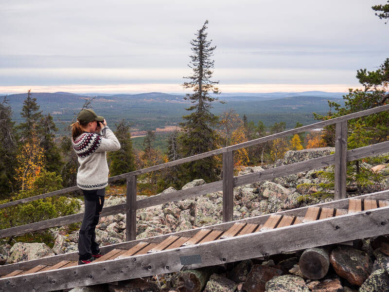 A person standing on wooden stairs along a stony hillside and photographing a view to forests and fell tops in autumn.