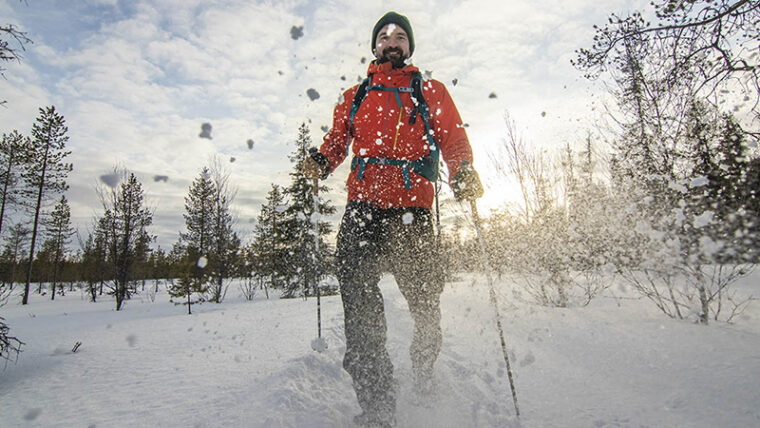 Hymyilevä ihminen kulkee sauvojen kanssa lumista polkua kohti kuvaajaa, lumi pöllyää ja aurinko paistaa. Taustalla kasvaa puita harvakseltaan.