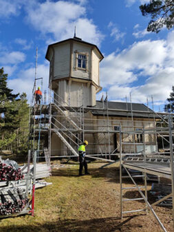 Ett gammalt trähus med torn. Omkring huset byggnadsställningar och på ställningarna en människa med hjälm och på marken en annan arbetare.