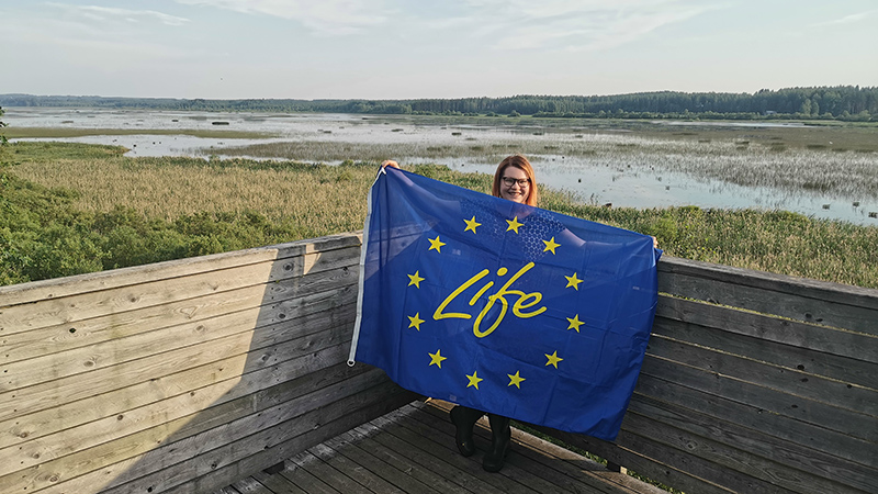 A smiling person standing on a birdwatching tower, holding a big flag wiht the word Life and EU stars. In the background a shallow lake with reeds.