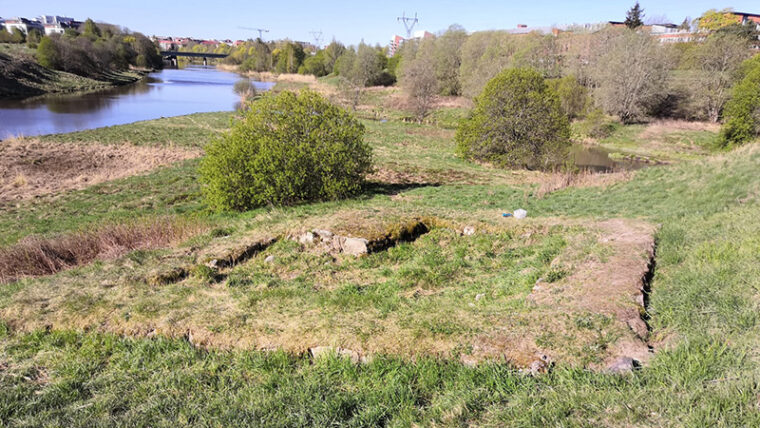 Nelikulmainen, matala ja ruohottunut kivirakenne joen rinteessä, taustalla kaupungin taloja.