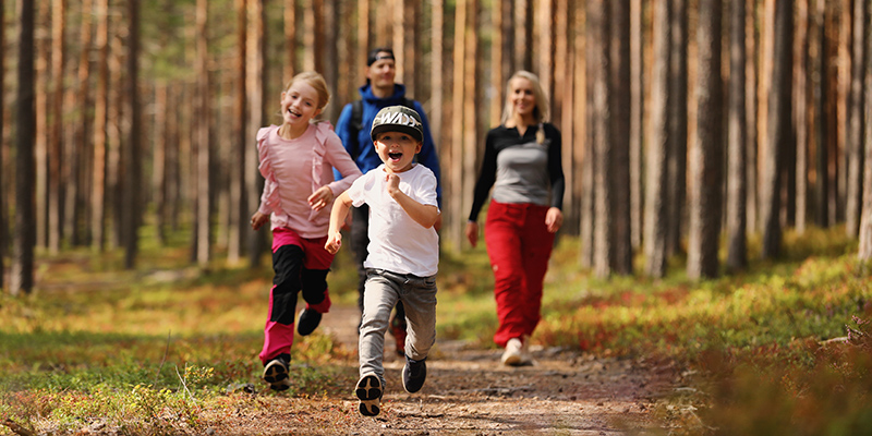 Kaksi lasta juoksee nauraen kohti kameraa metsäpolulla, takana kaksi aikusta ja mäntymetsää.