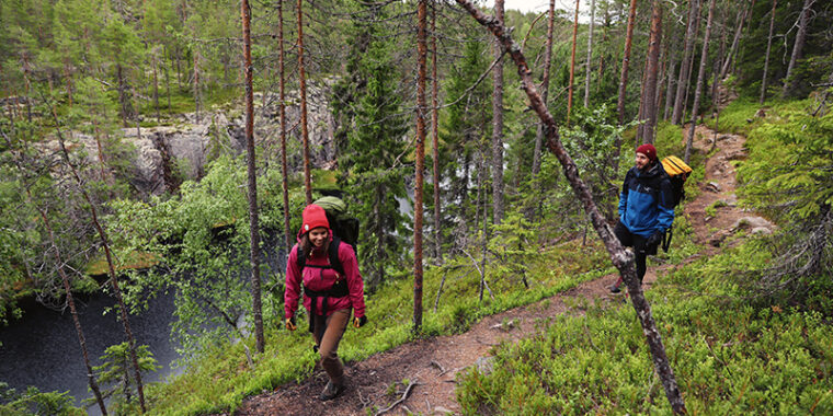 Kaksi ihmistä kulkee hymyillen metsäpolulla rinkat selässä, vieressä syvä rotko, jossa vettä.